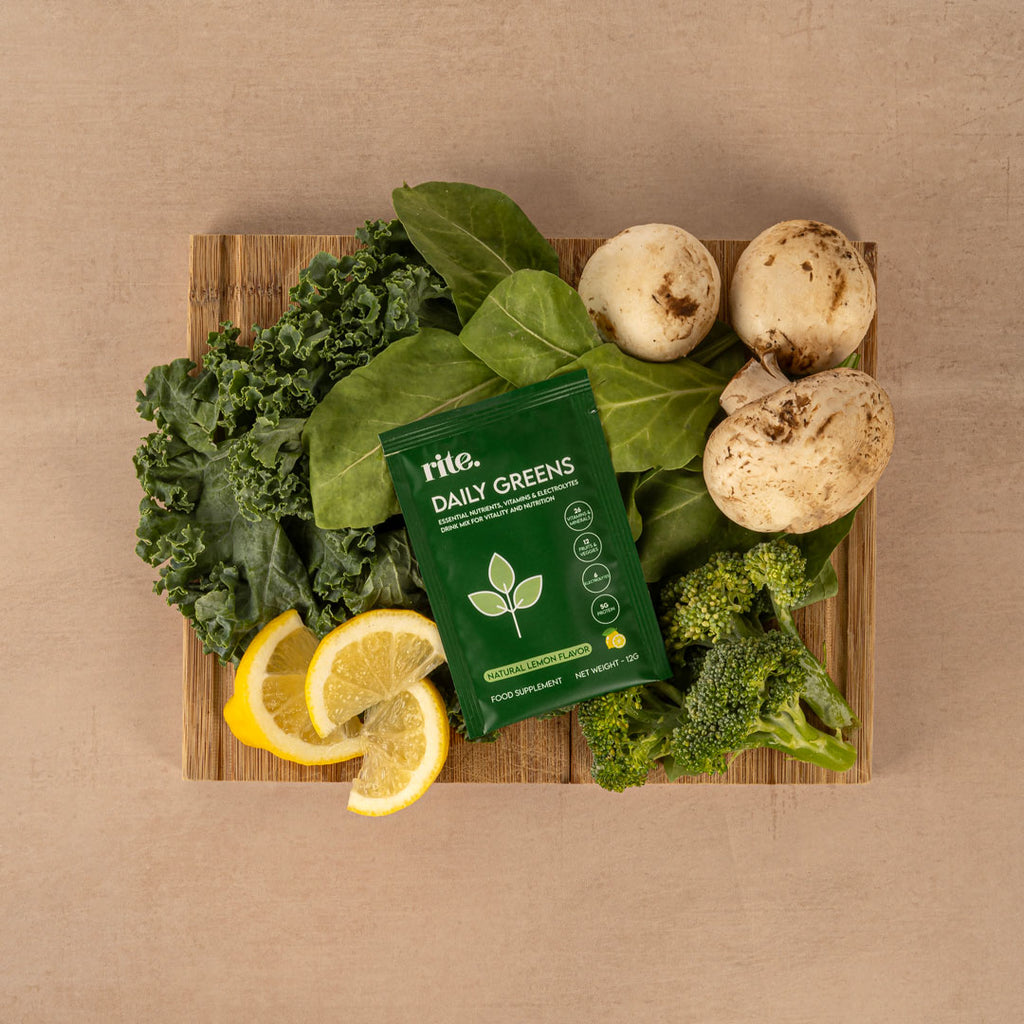 A green powder packet labeled "Rite Daily Greens" is placed on a wooden board surrounded by fresh vegetables