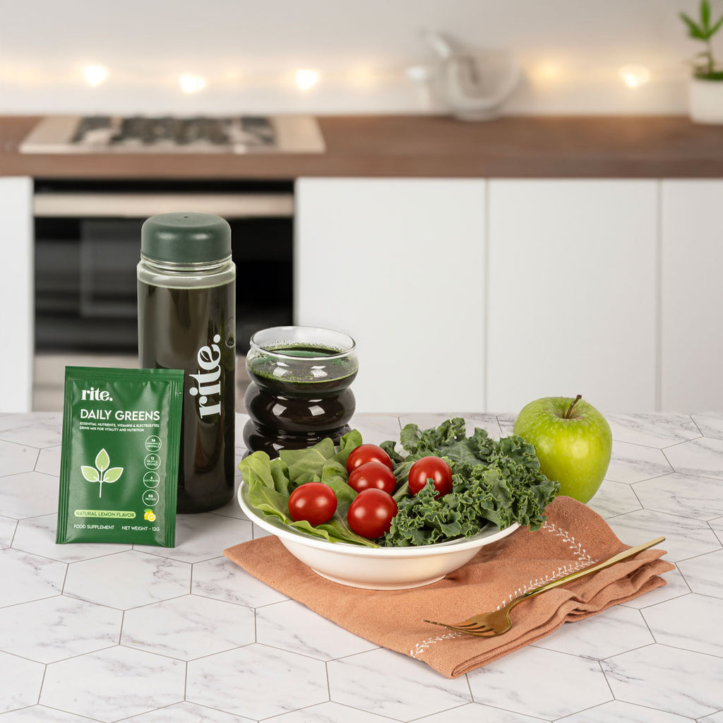 A green drink in a glass, a green powder packet labeled "Rite Daily Greens," a water bottle, and a bowl of cherry tomatoes and kale on a kitchen counter.