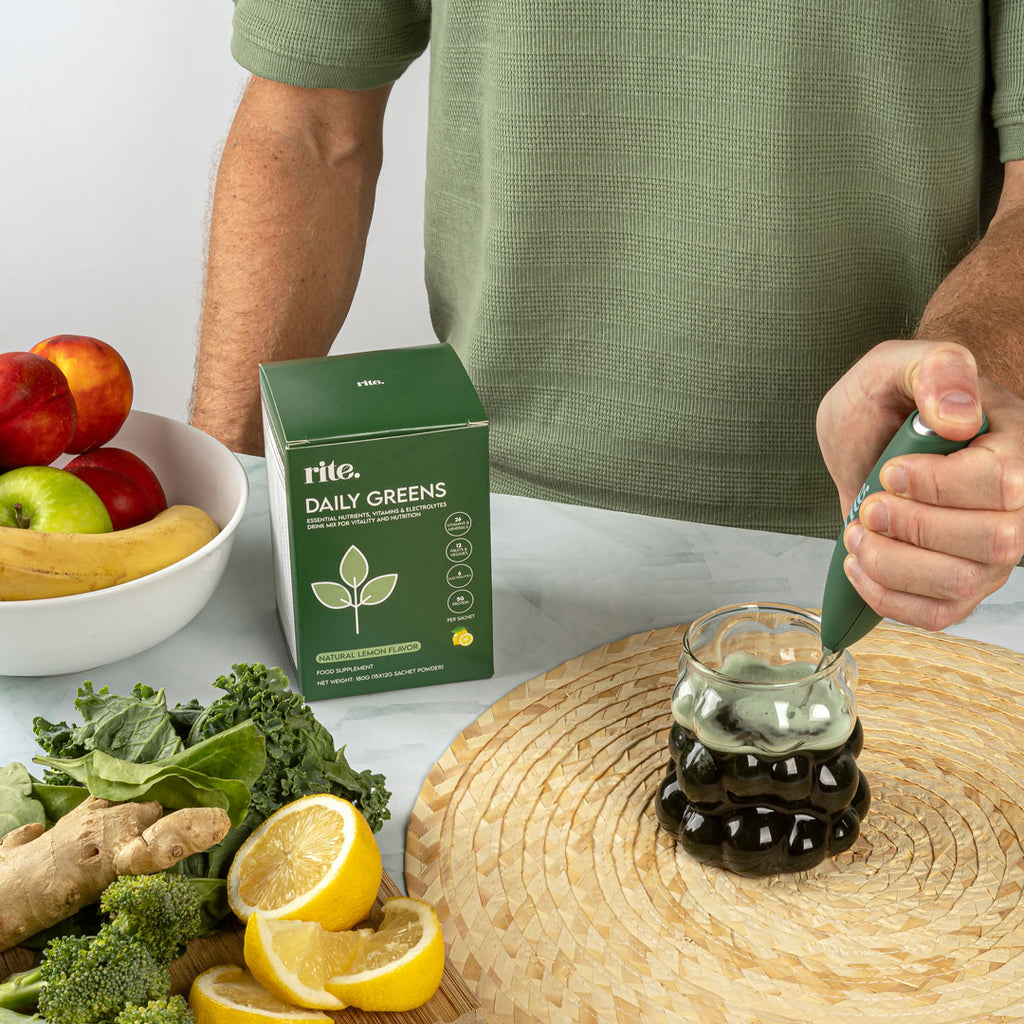 A man is using a blender to mix a green powder drink in a glass. The ingredients include spinach, ginger, lemon, and a box of "Rite Daily Greens".