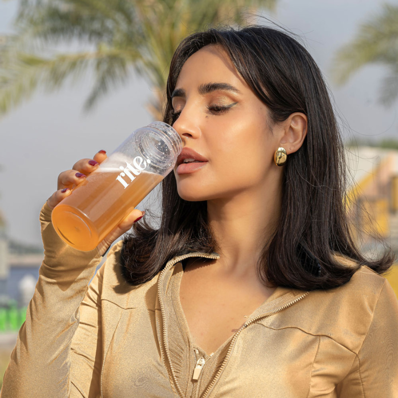 A woman in a beige jacket drinks an orange beverage from a clear bottle labeled "rite," standing outdoors with palm trees in the background.