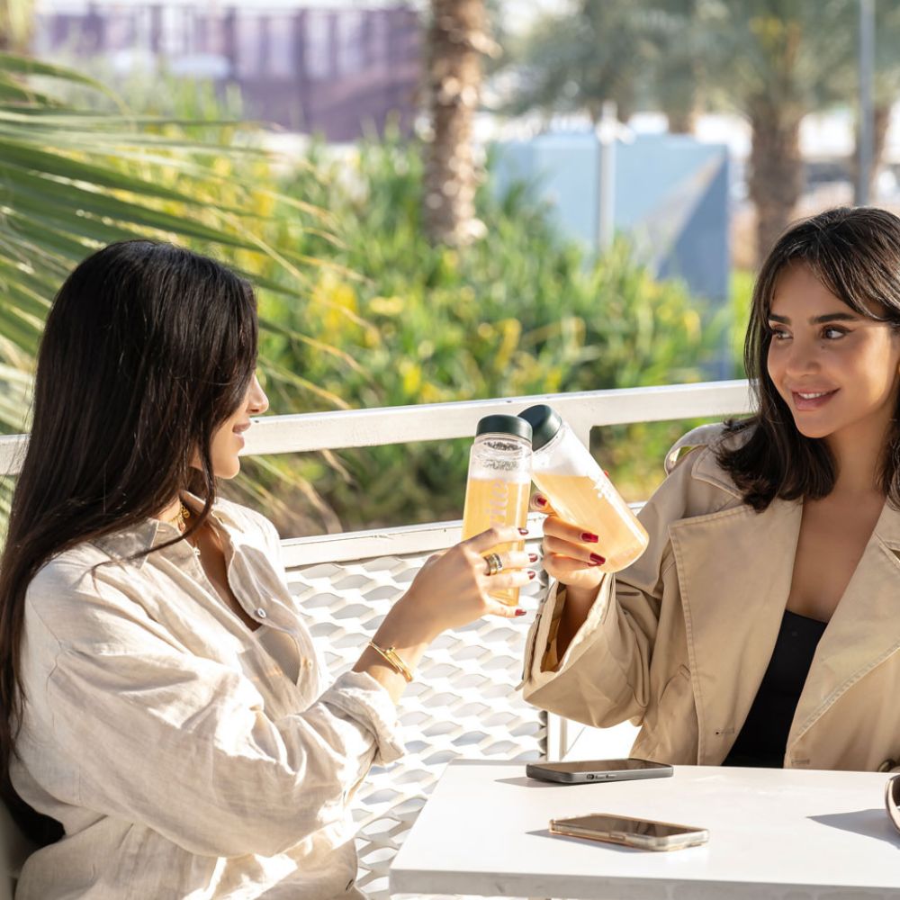 Two glasses of GUT HEALTH drinks with a light orange hue clinked together outdoors, highlighting a refreshing wellness beverage.