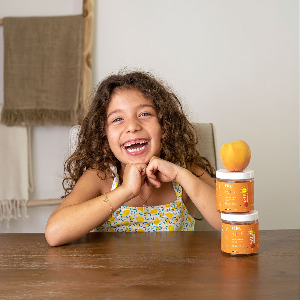 A girl happily sits at a table with a jar of KIDS gummies, smiling. The image promotes sugar-free multivitamin gummies for kids. 