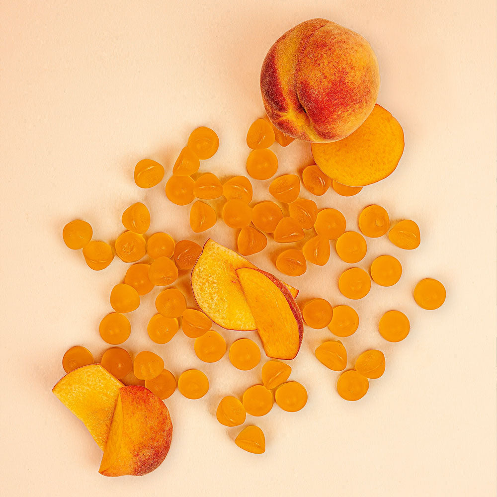 A pile of peaches and peach-colored IMMUNITY gummies  and slices sits on a light colored wooden table.
